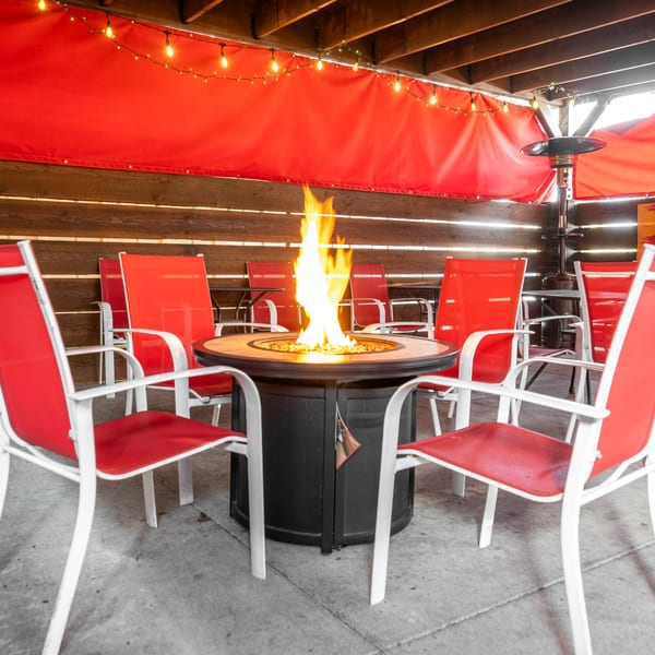 Outdoor patio with red chairs surrounding a fire pit, under string lights and red awnings.