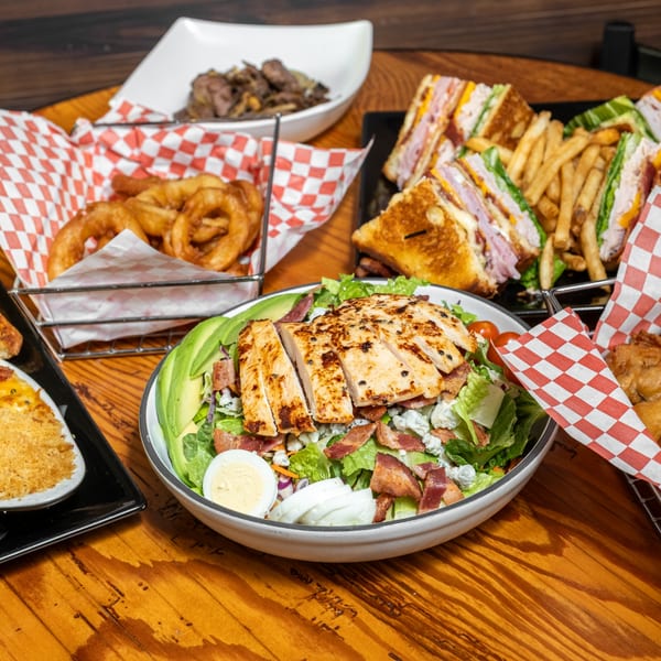 A grilled chicken salad, onion rings, club sandwiches, and other dishes on a wooden table.