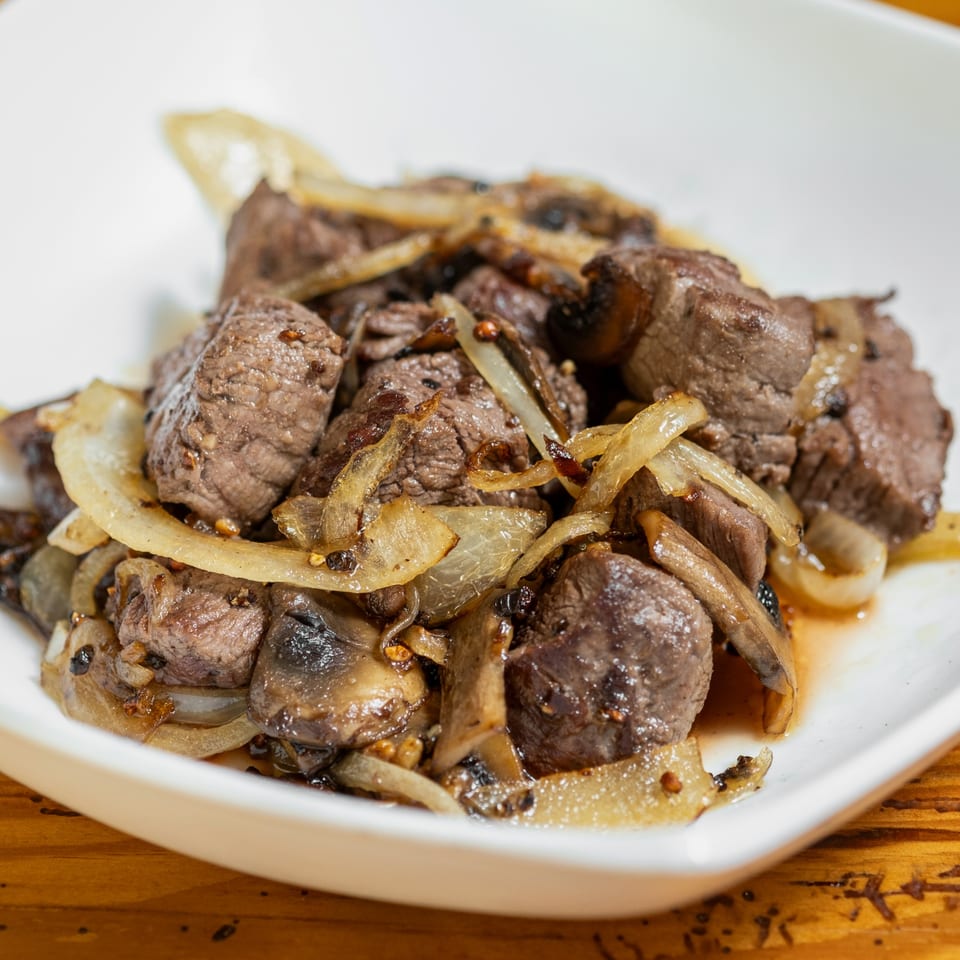 Close-up of steak bites with sautéed onions and mushrooms in a white bowl.