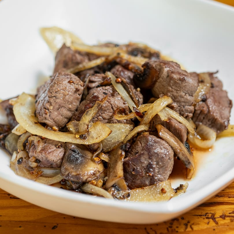 Close-up of steak bites with sautéed onions and mushrooms in a white bowl.