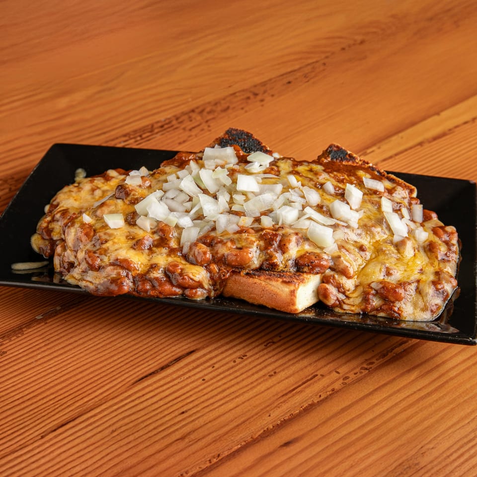 Dea's In & Out chili cheese toast, topped with diced white onions, on a black plate.
